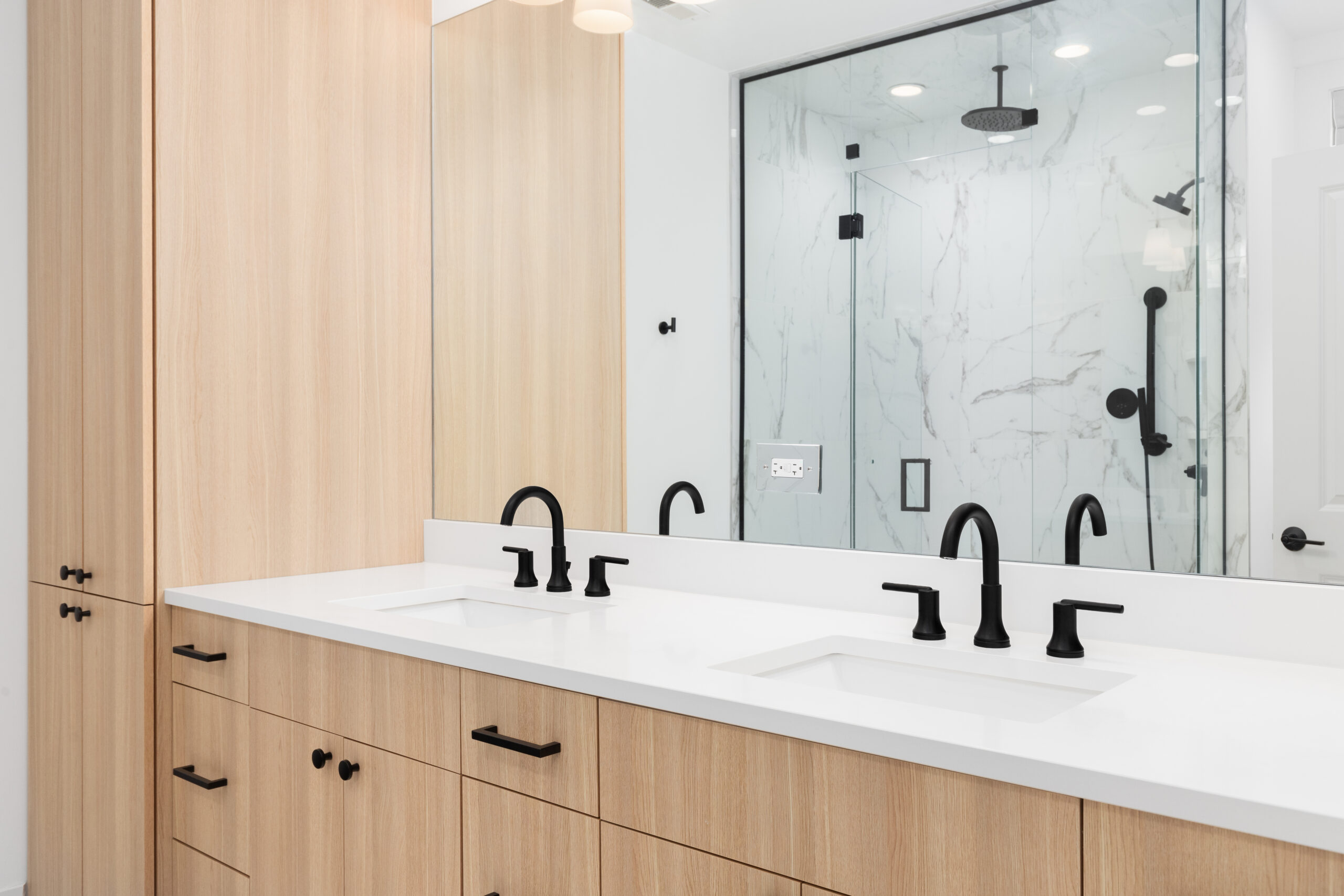 A beautiful bathroom detail with a wood cabinet, white marble countertop, black faucets, and a view of the walk-in shower through the mirror.